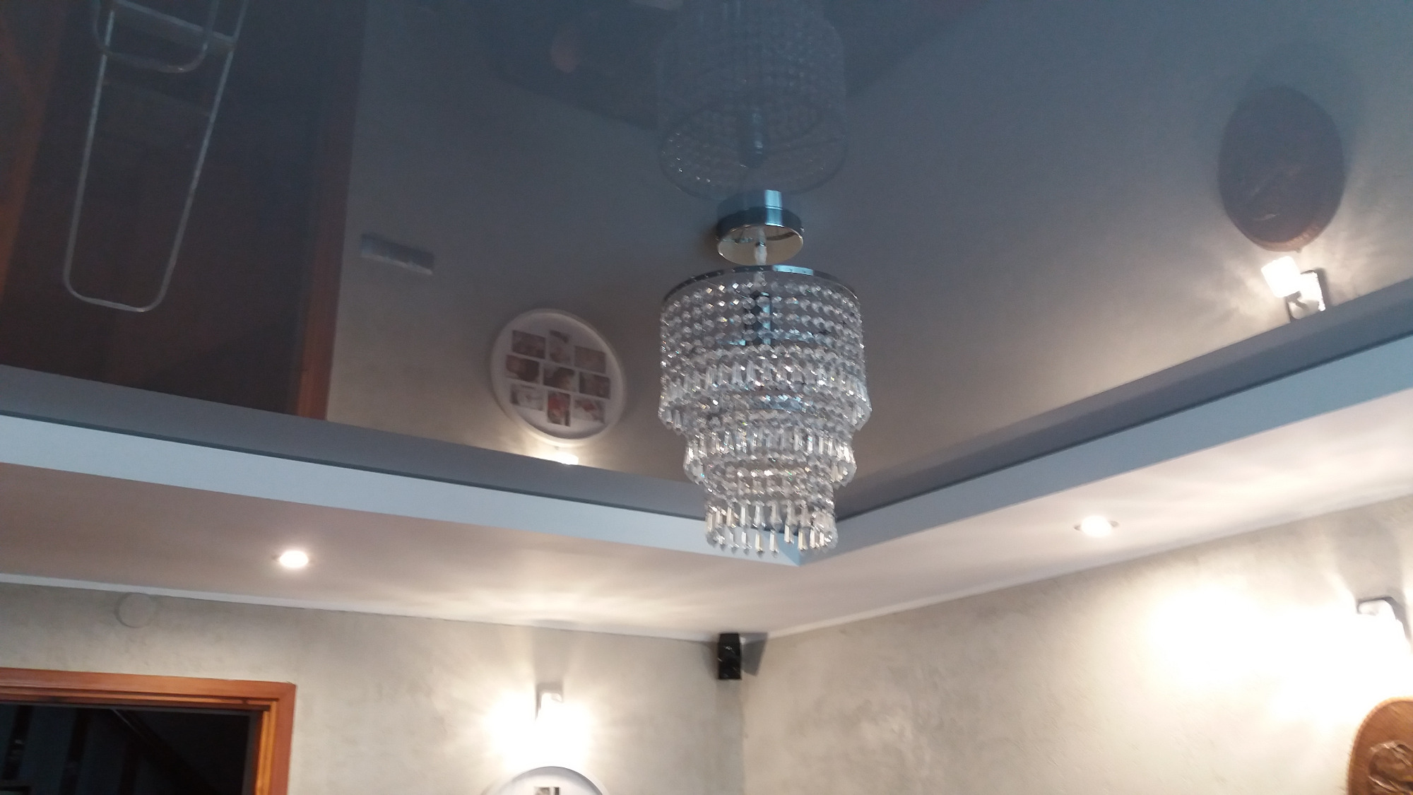 Błyszczący, kryształowy żyrandol wielopoziomowy zawieszony na błyszczącym, ciemnym suficie napinanym w wykończonym wnętrzu, z widocznymi elementami dekoracyjnymi na ścianach i oświetleniem punktowym.