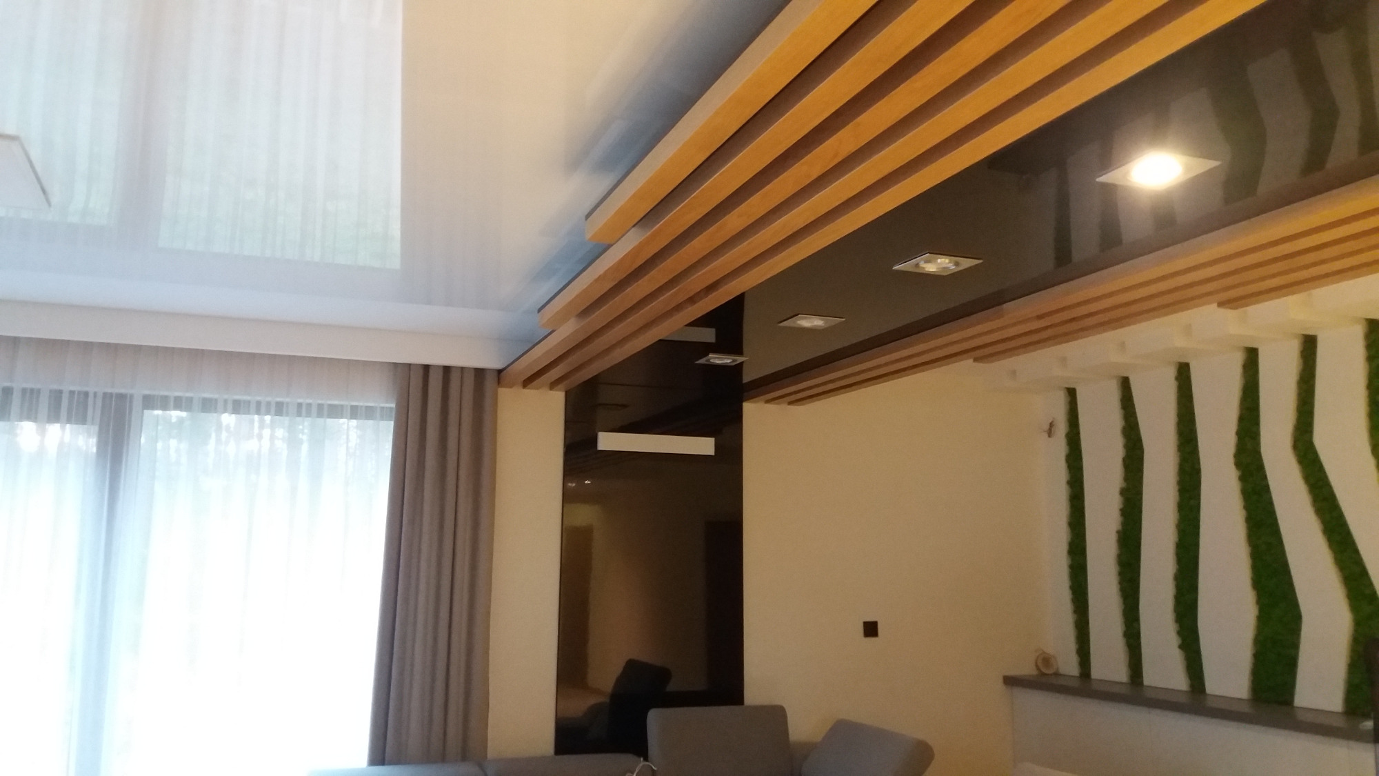 Nowoczesny salon z białym, błyszczącym sufitem napinanym, dekoracyjnymi drewnianymi listwami i podłużnym panelem z czarnego szkła na ścianie, oświetlonymi punktowo