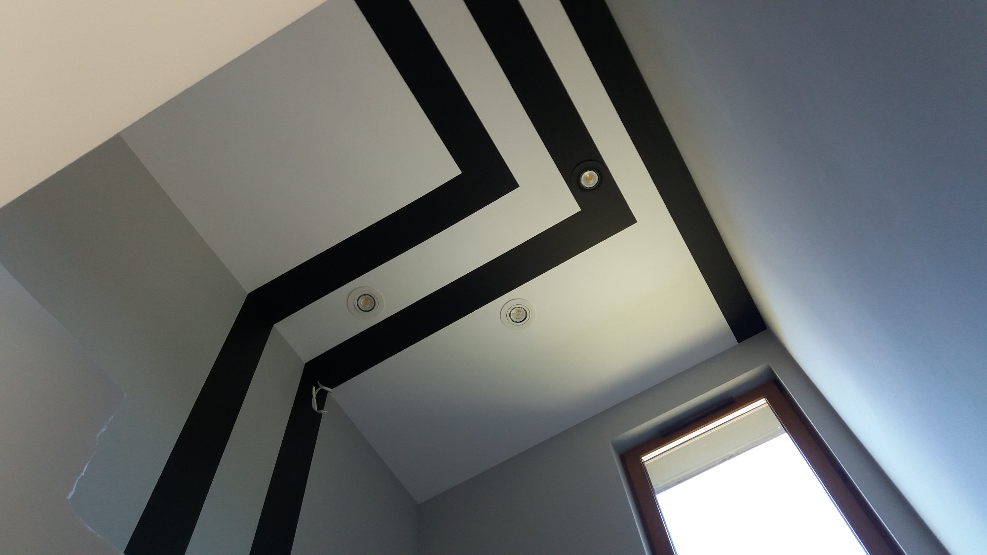 Biały sufit z czarnymi pasami tworzącymi geometryczny wzór, oświetlony trzema punktowymi lampami LED i fragmentem okna w prawym dolnym rogu.