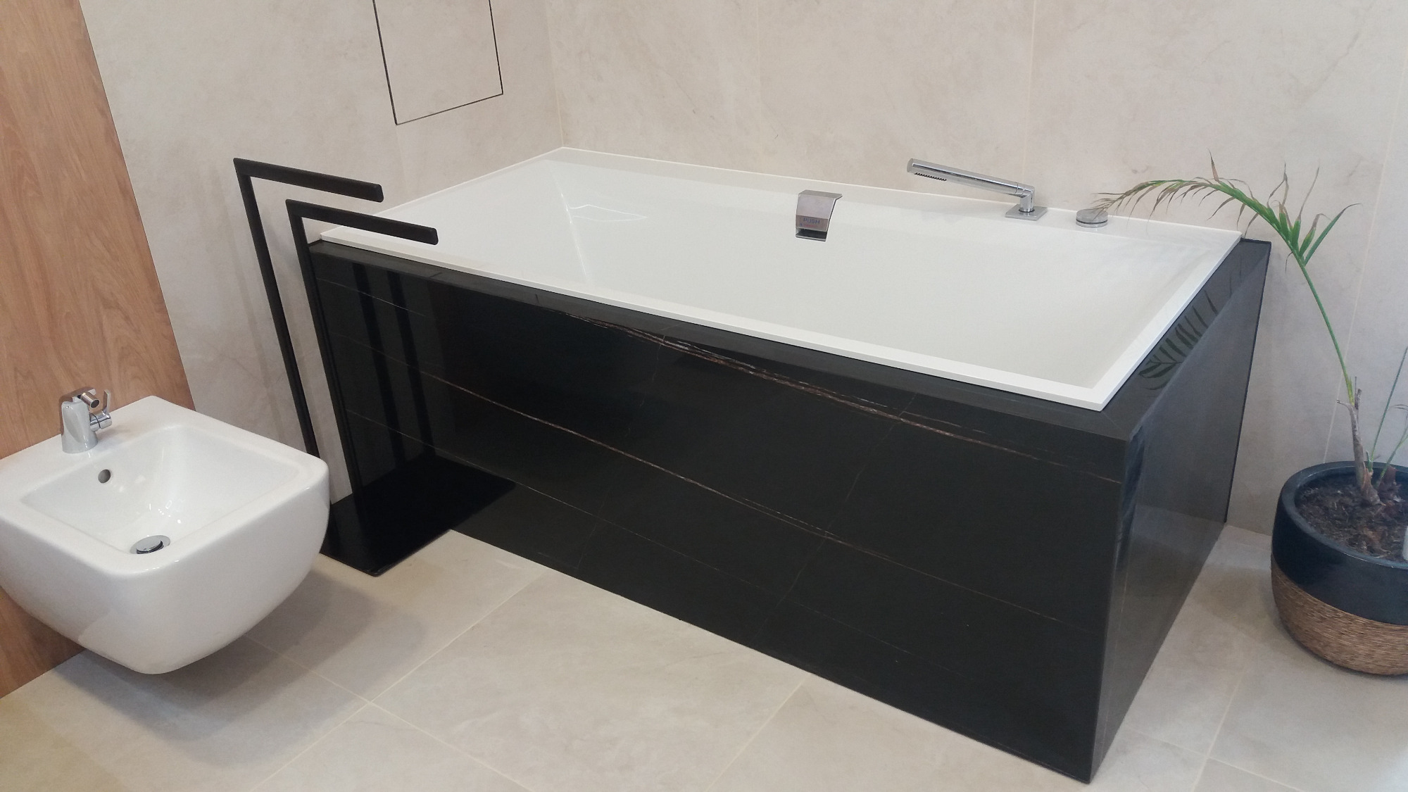 Nowoczesna łazienka z białą wanną obudowaną czarnym marmurem z brązowymi żyłkami, obok bidet i dekoracyjna roślina w czarnej doniczce.