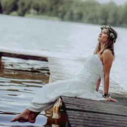 Kobieta w białej sukni i wianku na głowie siedzi na drewnianym pomoście nad jeziorem, spoglądając w górę.