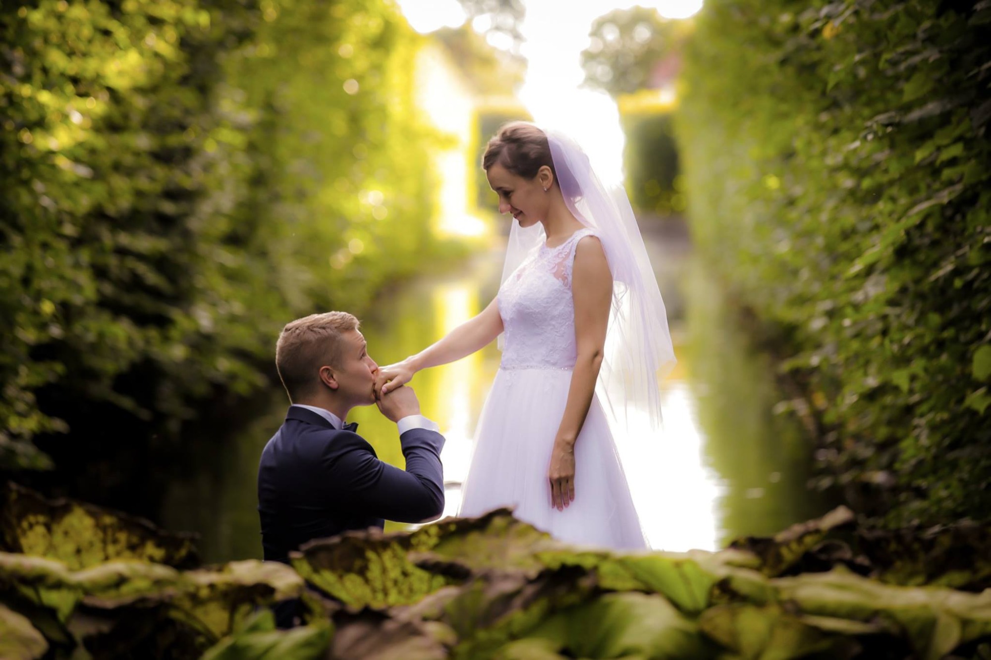Para młoda w romantycznej scenerii: Pan młody całuje dłoń panny młodej na tle zieleni.