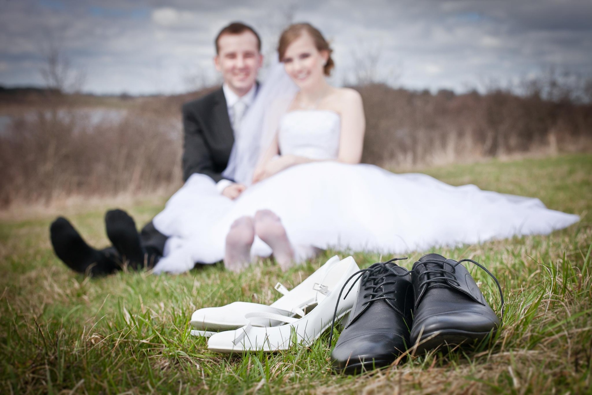Para młoda siedzi na trawie w tle, na pierwszym planie buty ślubne: białe damskie i czarne męskie sznurowane.