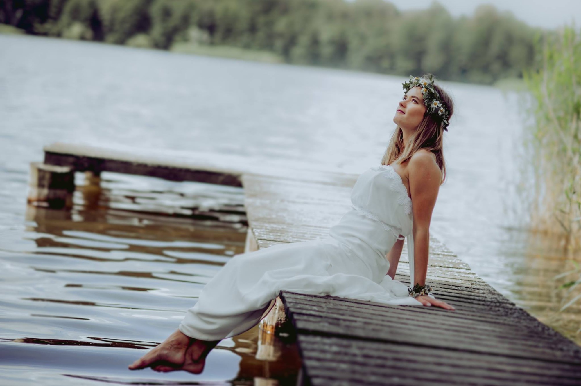 Kobieta w białej sukni i wianku na głowie siedzi na drewnianym pomoście nad jeziorem, spoglądając w górę.