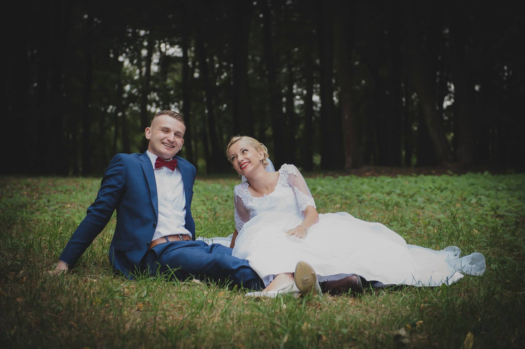 Uśmiechnięta para młoda w plenerze: on w granatowym garniturze z czerwoną muchą, ona w białej sukni ślubnej z koronkowymi rękawami, siedzą na trawie z lasem w tle.