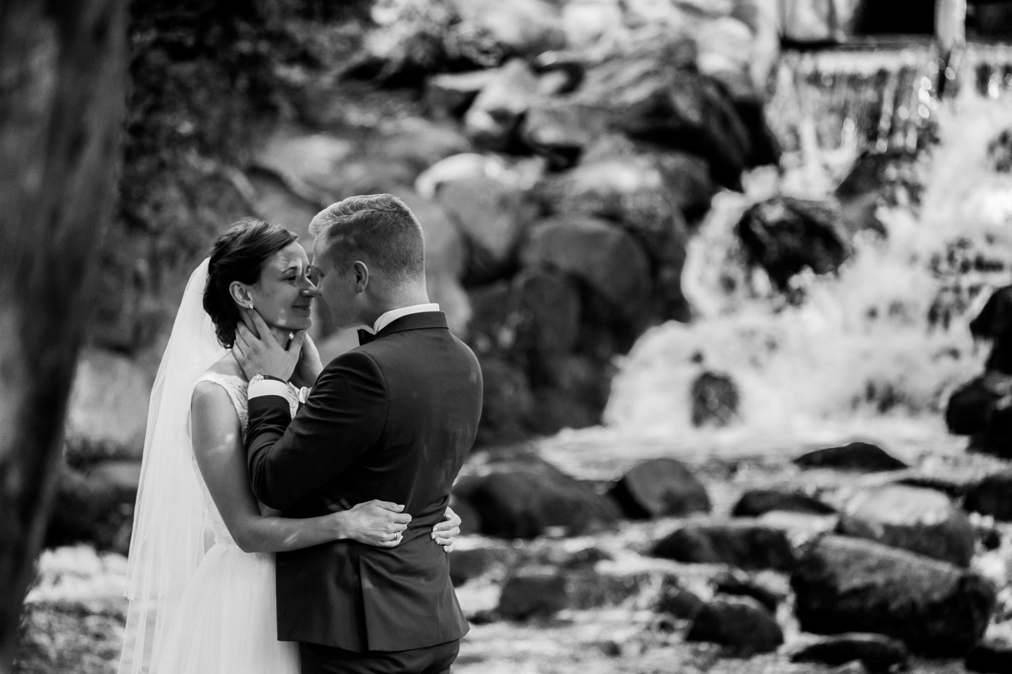 Czarno-białe zdjęcie pary młodej, obejmującej się na tle wodospadu w parku, panna młoda w welonie i sukni ślubnej, pan młody w ciemnym garniturze z muszką.