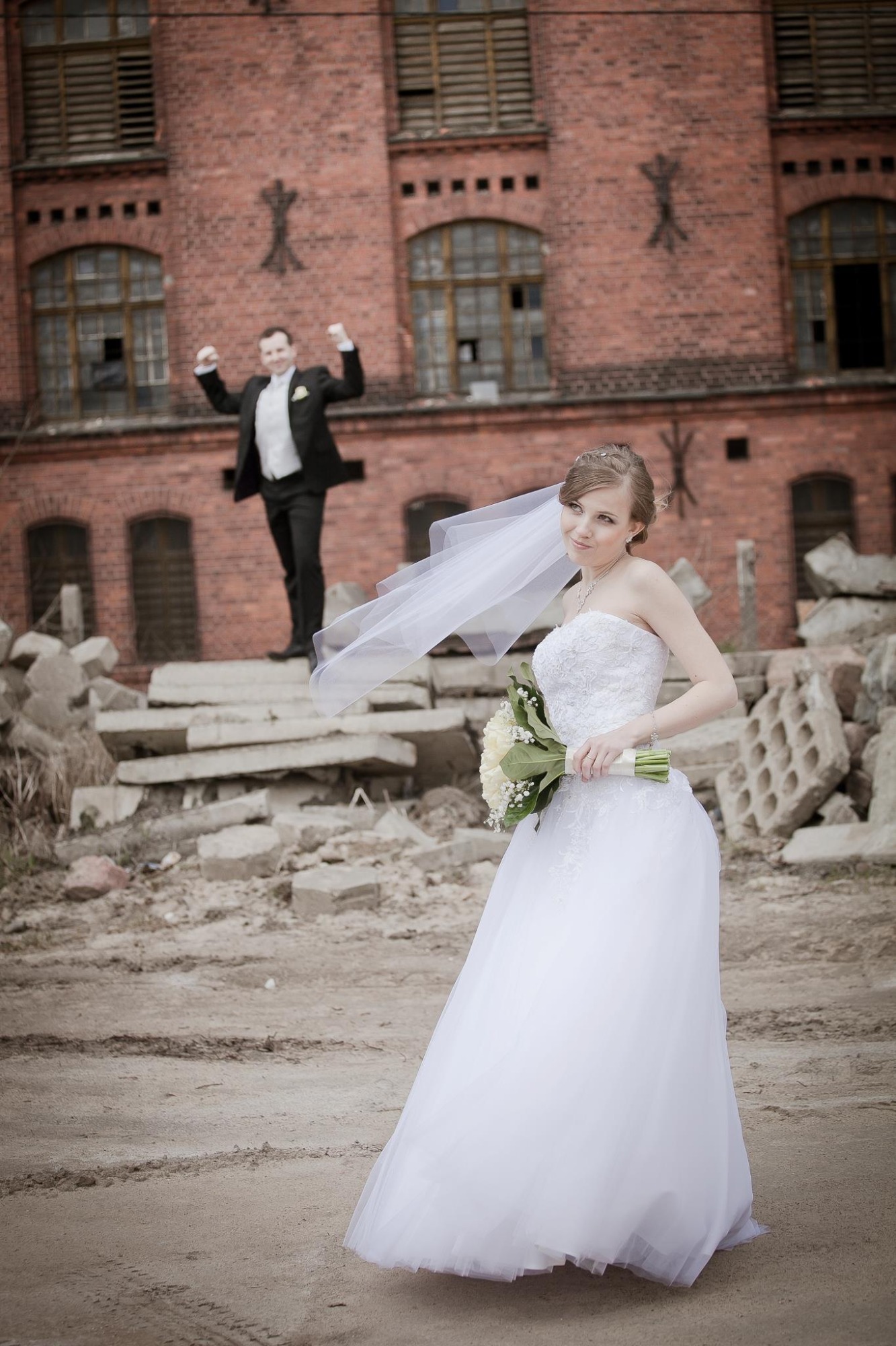Panna młoda w białej sukni ślubnej trzyma bukiet kwiatów na tle ceglanego budynku i gruzowiska, z uśmiechniętym panem młodym w tle z podniesionymi rękami.