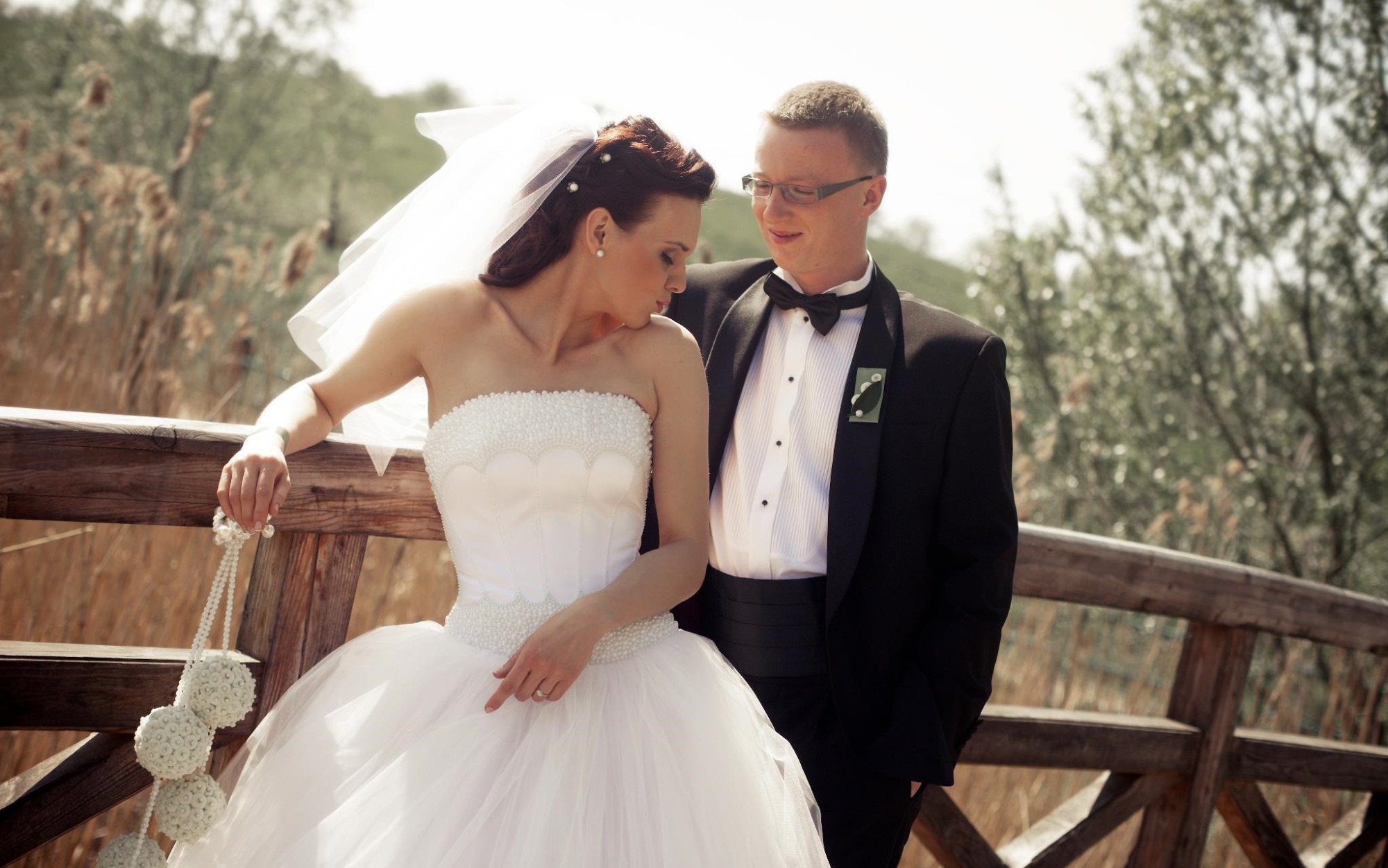 Para młoda w strojach ślubnych, panna młoda w sukni bez ramiączek z koralikami, trzyma dekorację z białych kul, pan młody w czarnym smokingu z ozdobną przypinką, opierają się o drewnianą barierkę...