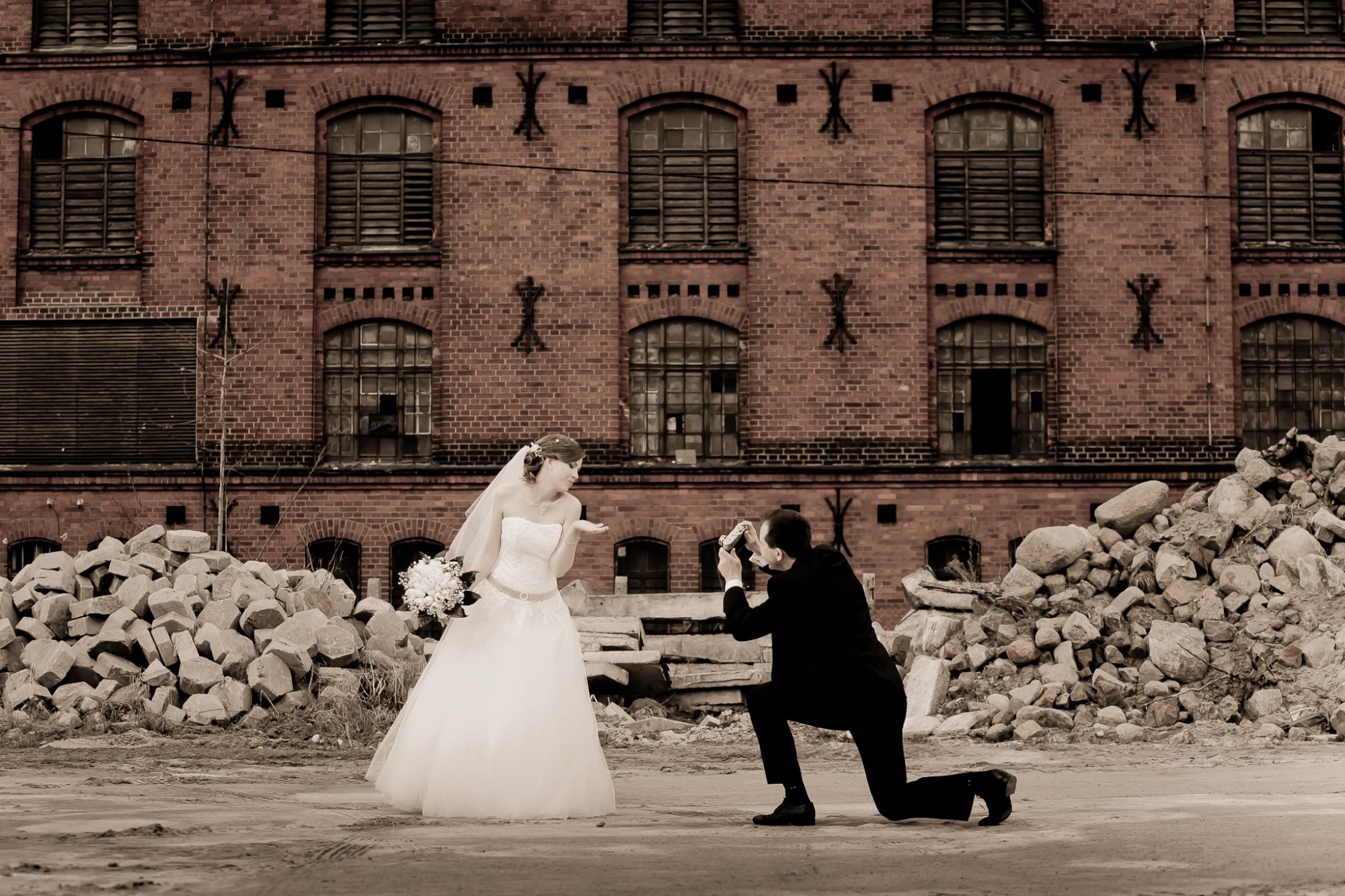 Panna młoda w białej sukni z bukietem pozuje do zdjęcia robionego przez fotografa klęczącego na ziemi, w tle ceglana ściana budynku i sterta kamieni.