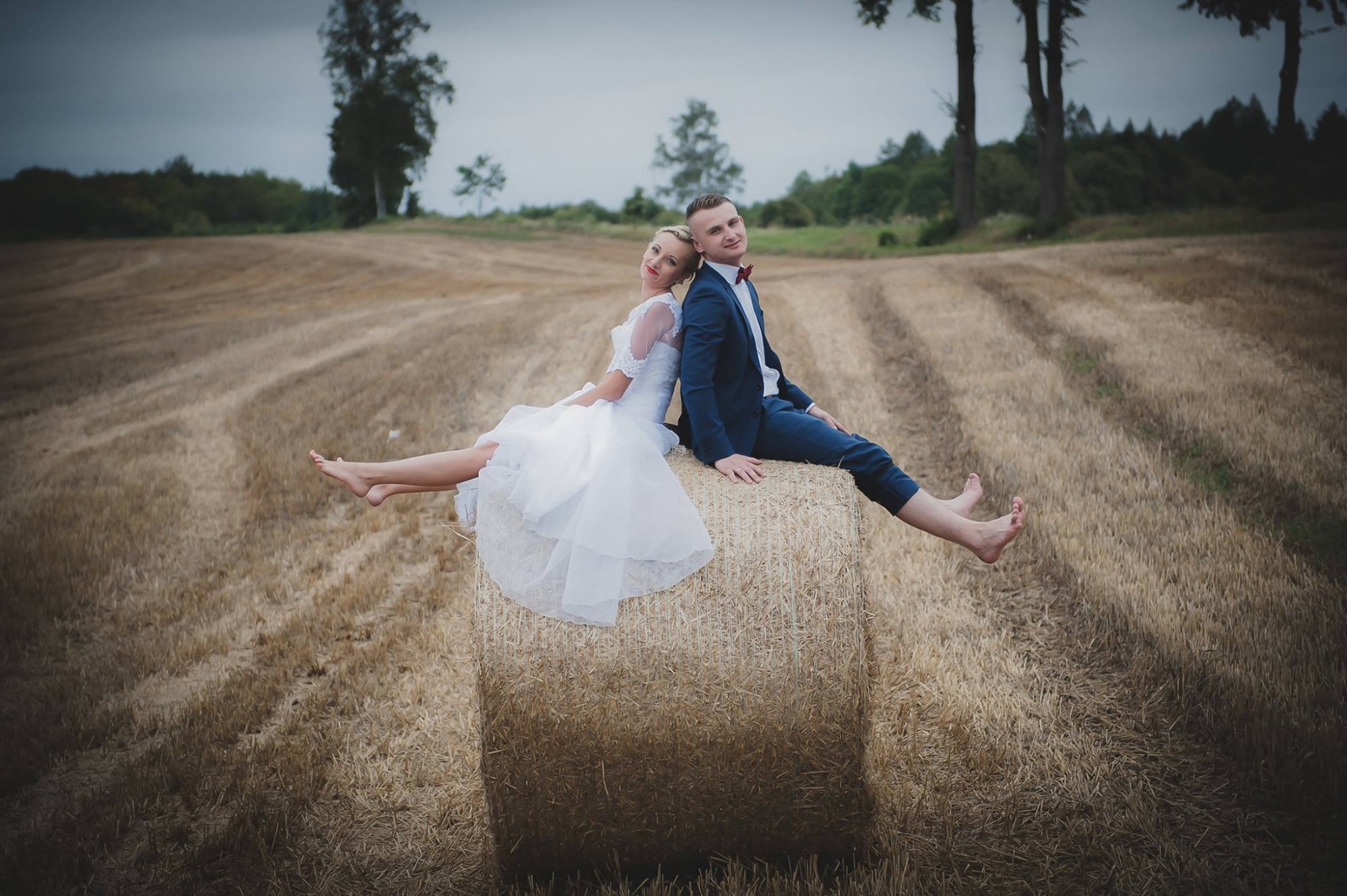 Para młoda, boso, siedzi plecami do siebie na belocie słomy na polu w pochmurny dzień.