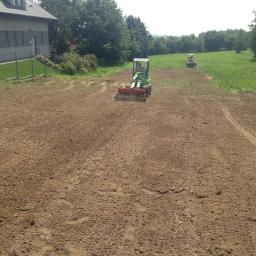 Piokop - Dwa traktory wyrównują teren na polu w Warszawie, przygotowując glebę pod uprawę. W tle widoczny budynek i drzewa.