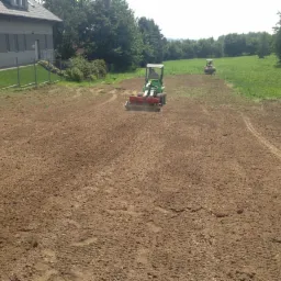Dwa traktory wyrównują teren na polu w Warszawie, przygotowując glebę pod uprawę. W tle widoczny budynek i drzewa.