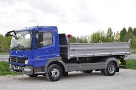 Niebieski wywrotka Mercedes-Benz Atego z aluminiową skrzynią ładunkową, stojąca na żwirowym podłożu, widoczna z boku, w tle drzewa i droga.