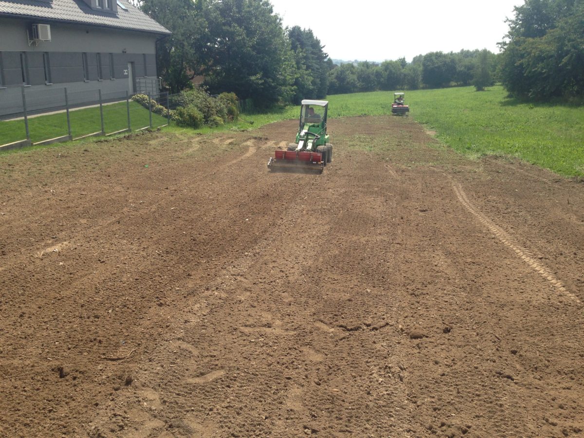 Dwa traktory wyrównują teren na polu w Warszawie, przygotowując glebę pod uprawę. W tle widoczny budynek i drzewa.