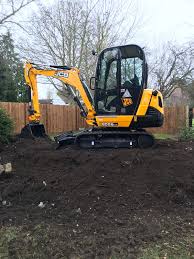 Żółta mini koparka JCB na nierównym, świeżo przekopanym terenie z widocznym ogrodzeniem drewnianym w tle.