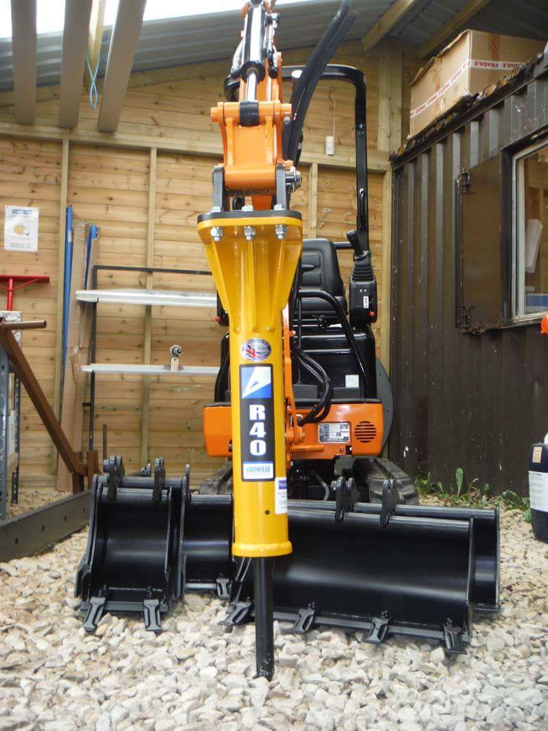 Żółto-pomarańczowa mini koparka z zamontowanym młotem hydraulicznym R40 i dwiema czarnymi łyżkami stoi na żwirowym podłożu przed drewnianą ścianą i kontenerem.