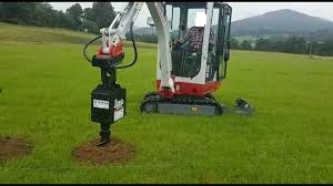 Mini koparka z wiertnicą hydrauliczną wiercąca otwór w trawiastym terenie, widoczne wywiercone otwory, w tle wzgórza i drzewa.