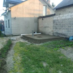 Świeżo wylana, gładka betonowa wylewka fundamentowa w Poniatowej, z widocznymi rurami i włazem kanalizacyjnym, otoczona ziemią i roślinnością, przylegająca do ściany budynku z pustaków.