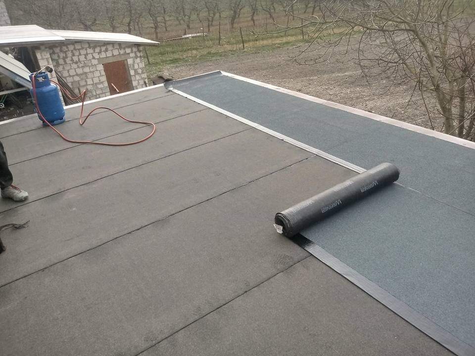 Widok z góry na płaski dach pokryty papą termozgrzewalną. Część dachu już pokryta, obok leży rolka papy. W tle budynek gospodarczy i butla z gazem.