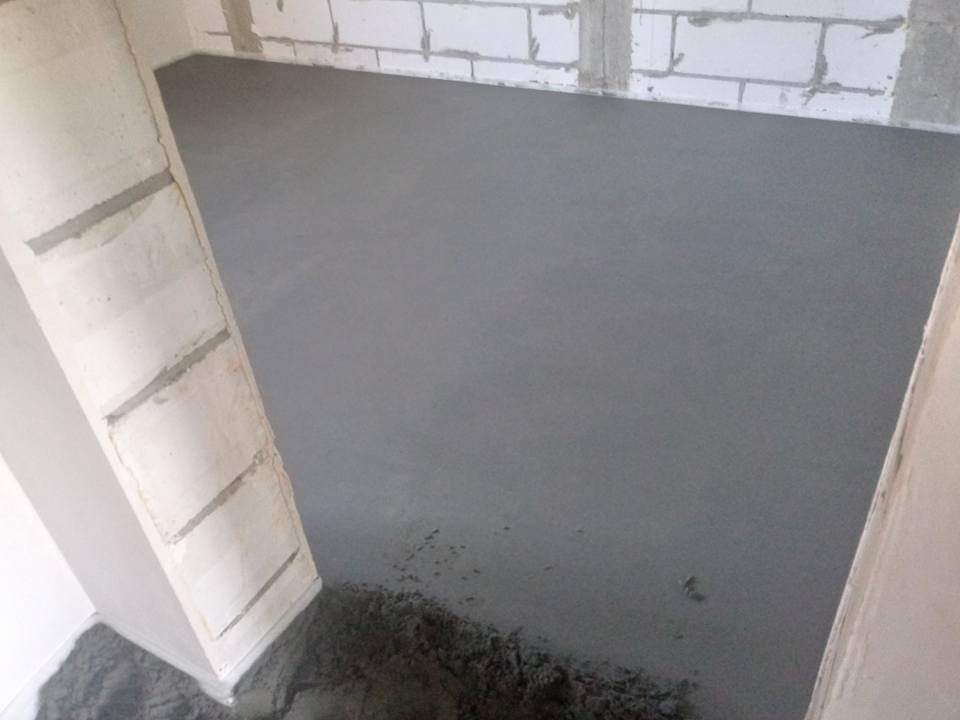Świeżo wylana, gładka, szara posadzka w pomieszczeniu z surowymi ścianami z betonu komórkowego, widoczne niewielkie pozostałości materiału w rogu.