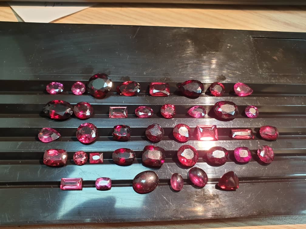 Różnorodny zbiór oszlifowanych kamieni szlachetnych w odcieniach czerwieni i różu, ułożonych na czarnej, rowkowanej powierzchni, o różnych kształtach i rozmiarach, z widocznymi refleksami świetlnymi.