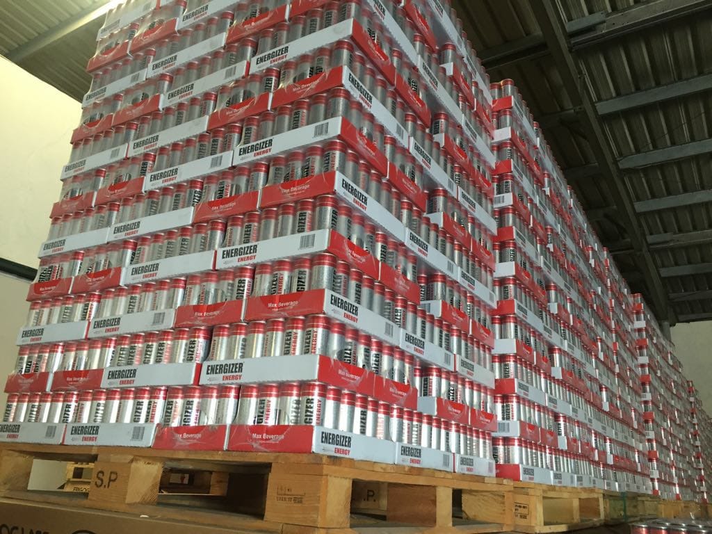 Wysoki stos palet z napojami energetycznymi Energizer w srebrnych puszkach z czerwonymi elementami, składowanych w magazynie z widoczną konstrukcją dachu.