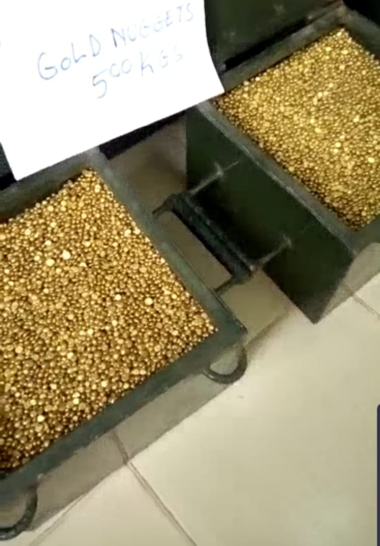 Dwa metalowe pojemniki wypełnione złotymi bryłkami, obok nich kartka z odręcznym napisem 'Gold Nuggets 500 kgs'.