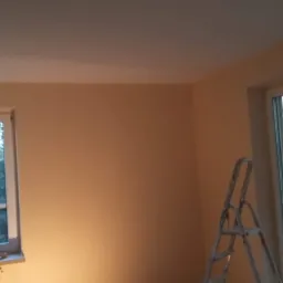 Świeżo pomalowany pokój na delikatny odcień brzoskwini, widoczna drabina malarska przy oknie balkonowym i narzędzia malarskie na parapecie pod oknem.