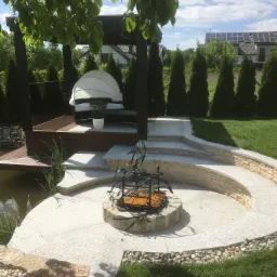 Ogród z okrągłym paleniskiem z kamienia, tarasem z zadaszeniem i leżanką, oraz oczkiem wodnym, otoczony tujami i zielonym trawnikiem w słoneczny dzień.