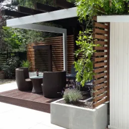 Nowoczesny taras z drewnianą podłogą, meblami rattanowymi pod biało-brązową pergolą, obok betonowa donica z lawendą i drewniana ażurowa ścianka.