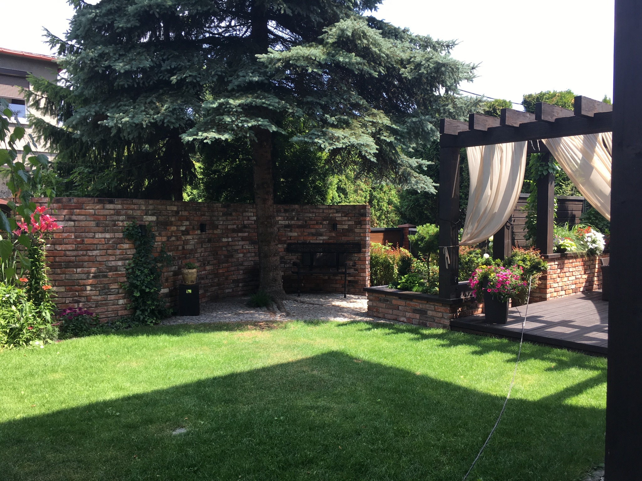 Zacieniony trawnik z widokiem na murowany grill, drewniany taras z pergolą i kwitnące kwiaty w donicach, otoczony bujną zielenią.