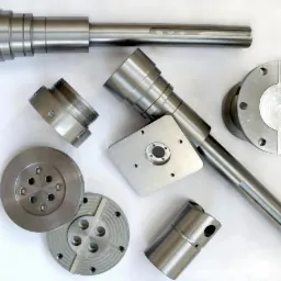 Zestaw precyzyjnie obrobionych, metalowych elementów o różnym kształcie, w tym wały, tarcze i tuleje, leżących obok suwmiarki na jasnym tle.