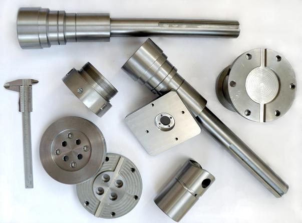 Zestaw precyzyjnie obrobionych, metalowych elementów o różnym kształcie, w tym wały, tarcze i tuleje, leżących obok suwmiarki na jasnym tle.