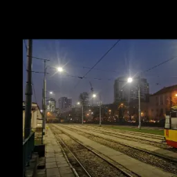 Szyny tramwajowe nocą oświetlone przez lampy uliczne, tramwaj częściowo widoczny po prawej stronie kadru, w tle budynki mieszkalne i drzewa.