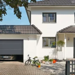 Biały dom jednorodzinny z szarą bramą garażową, na podjeździe rower, rośliny w doniczkach, a przed wejściem leży pies.