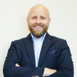 Uśmiechnięty mężczyzna z brodą, ubrany w niebieską marynarkę i koszulę, stoi na jasnym tle z założonymi rękami. Widoczny fragment poszetki w kieszonce.