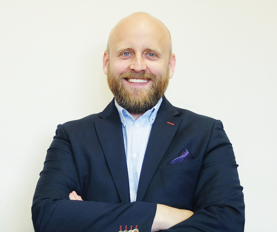 Uśmiechnięty mężczyzna z brodą, ubrany w niebieską marynarkę i koszulę, stoi na jasnym tle z założonymi rękami. Widoczny fragment poszetki w kieszonce.