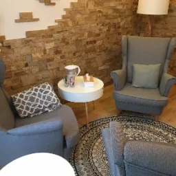 Przytulne wnętrze gabinetu z trzema szarymi fotelami typu uszak, okrągłym stolikiem z dzbankiem filtrującym i pudełkiem chusteczek, oraz dywanem w etniczne wzory na drewnianej podłodze, ściana...