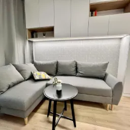 Szara sofa narożna z poduszkami w pokoju z białymi szafkami wiszącymi i geometryczną tapetą, mały czarny stolik kawowy z wazonem z białymi kwiatami.