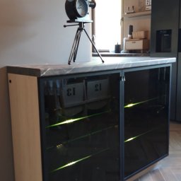 FHU ANMEBEL - Szafka RTV z przeszklonymi, podświetlanymi półkami, na blacie dekoracyjny model kamery filmowej, w tle widoczna kuchnia