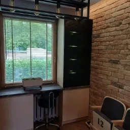 Kącik z biurkiem pod oknem, zabudowa z czarną witryną, ceglana ściana i drewniane krzesła kinowe z numerami 12 i 13.