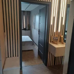 Nowoczesna aranżacja przedpokoju z drewnianymi lamelami na ścianach, siedziskiem z tapicerowanym wiekiem i otwartymi drzwiami do sypialni z parkietem w jodełkę.