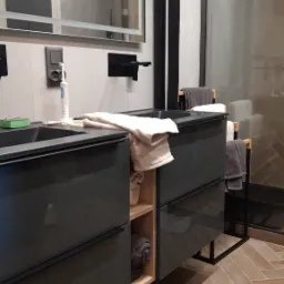 Nowoczesna szafka łazienkowa na wymiar z szufladami i otwartymi półkami na ręczniki, zrealizowana w Piotrkowie Trybunalskim, z drewnianą podłogą ułożoną w jodełkę.