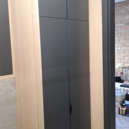 FHU ANMEBEL - Wysoka, grafitowa szafa w zabudowie z drewnianymi bokami, z minimalistycznymi, podłużnymi uchwytami, widoczna z boku, obok wejście do pokoju z ceglaną ścianą.