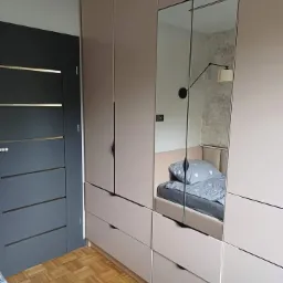 Jasna zabudowa meblowa z szafami, szufladami i lustrzanym frontem, odbijająca fragment sypialni z łóżkiem i lampką ścienną, na tle parkietu ułożonego w kwadraty oraz ciemnych drzwi z poziomymi...