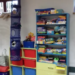 Regał z półkami i szufladami wypełniony grami planszowymi i puzzlami, obok regał z kolorowymi pojemnikami na zabawki, wiszący organizer siatkowy i pluszowy miś na parapecie.