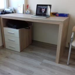 FHU ANMEBEL - Biurko z szufladami w odcieniach drewna i bieli, na blacie laptop z uruchomionym filmem edukacyjnym, obok pudełko rękawic nitrylowych, na podłodze panele imitujące drewno.