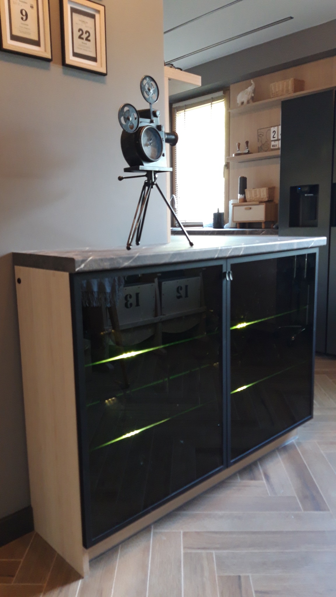 Szafka RTV z przeszklonymi, podświetlanymi półkami, na blacie dekoracyjny model kamery filmowej, w tle widoczna kuchnia