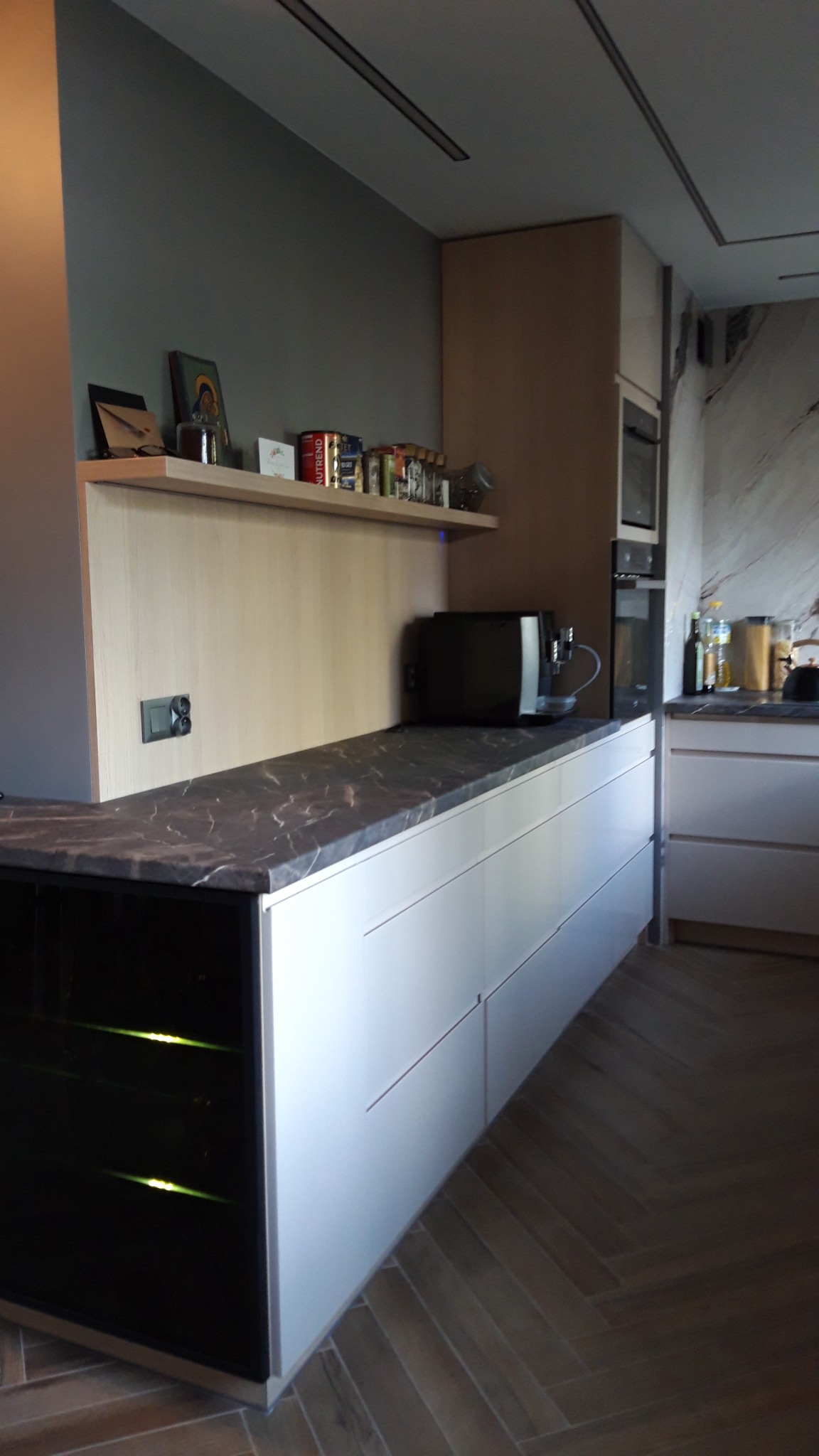 Nowoczesna kuchnia z białymi szafkami, blatem z ciemnego marmuru i szklaną witryną na wino, uzupełniona półką z dekoracjami i sprzętem AGD.