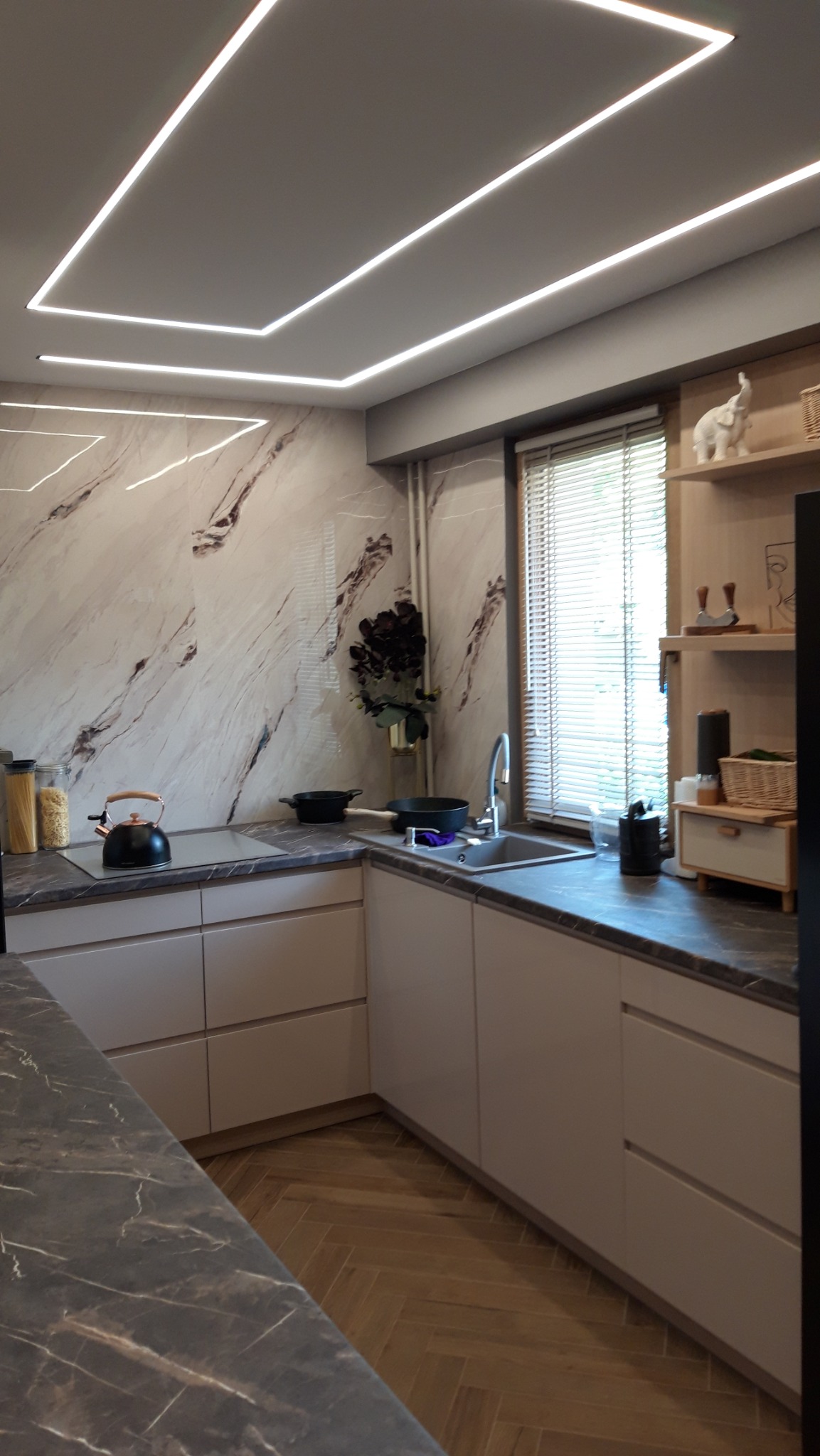 Nowoczesna kuchnia z beżowymi frontami, marmurową ścianą i podświetlanym sufitem, widok na zlew i część blatu z czajnikiem.